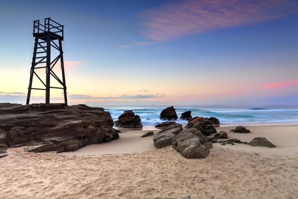 Wooden lookout tower on rocky outcrop overlooking redhed's sandy beach and ocean at sunset.

